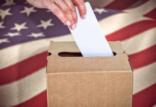 Person turning in a ballot into a cardboard voting box with the American flag in the background