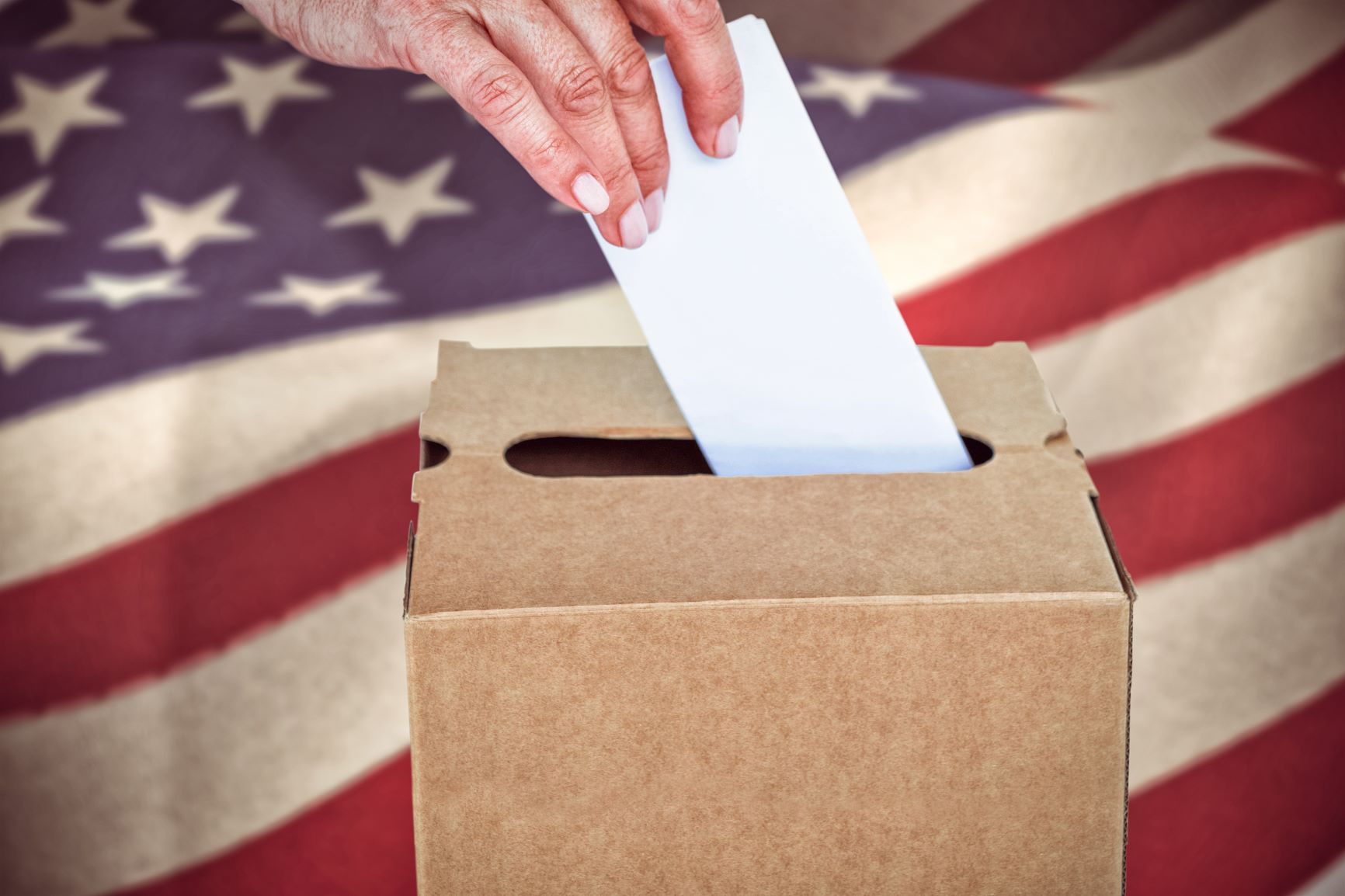 Person turning in a ballot into a cardboard voting box with the American flag in the background