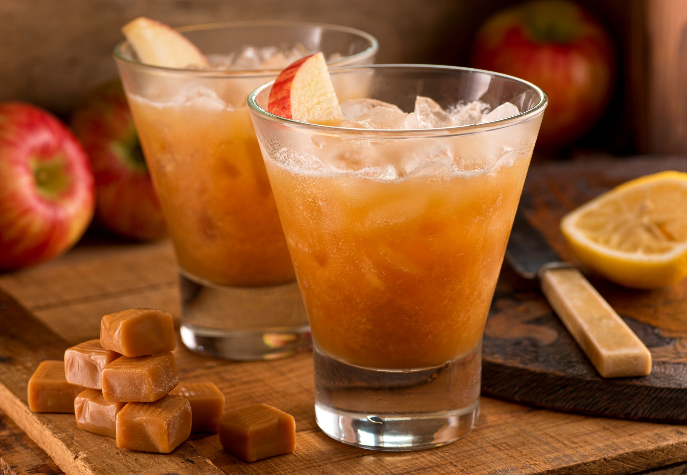 two glasses of fall caramel apple cocktails with apple slice garnishes on a wood table with caramel candies and apples.
