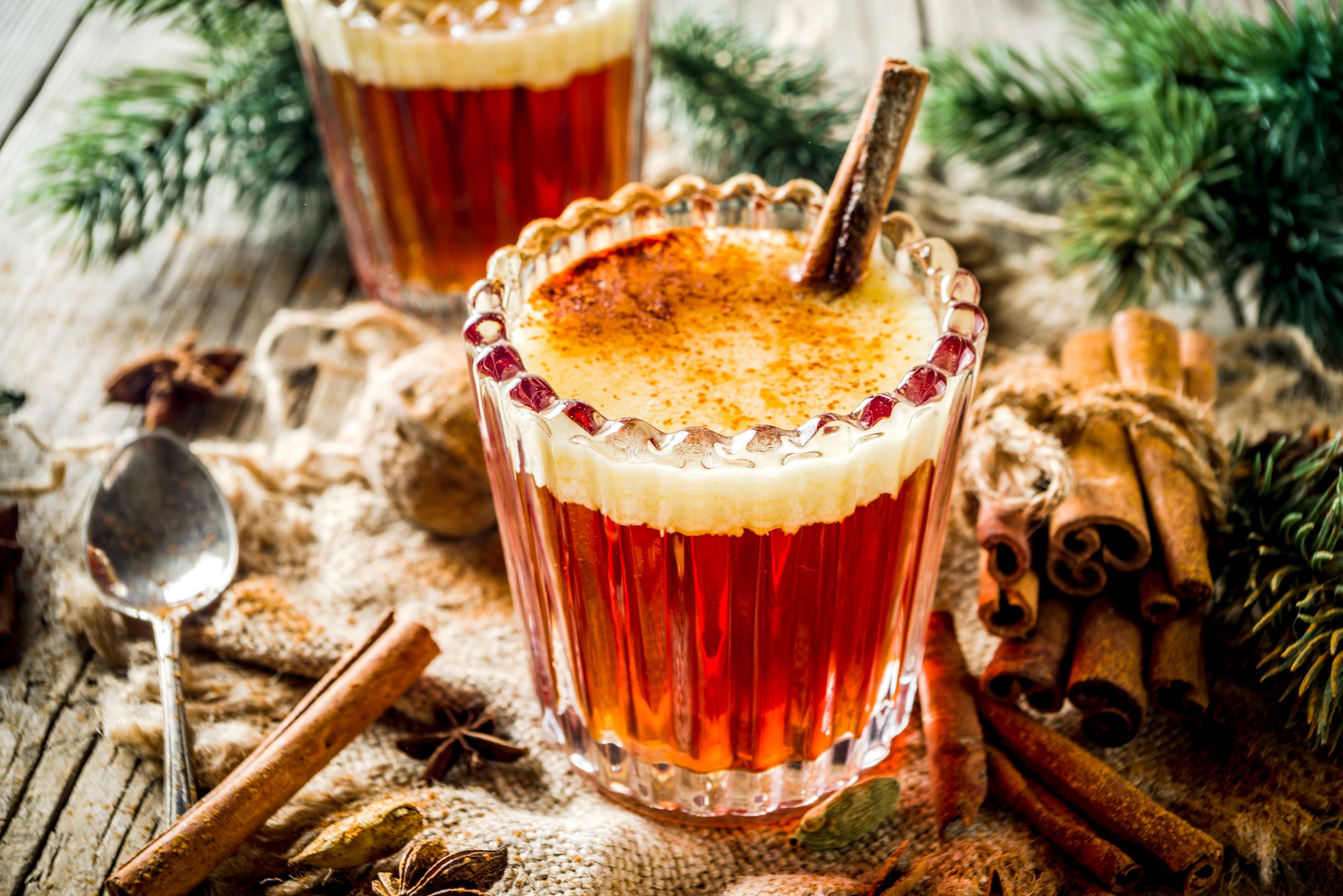 Glass of festive hot buttered rum on a table with cinnamon sticks, a spoon and sprigs of pine tree