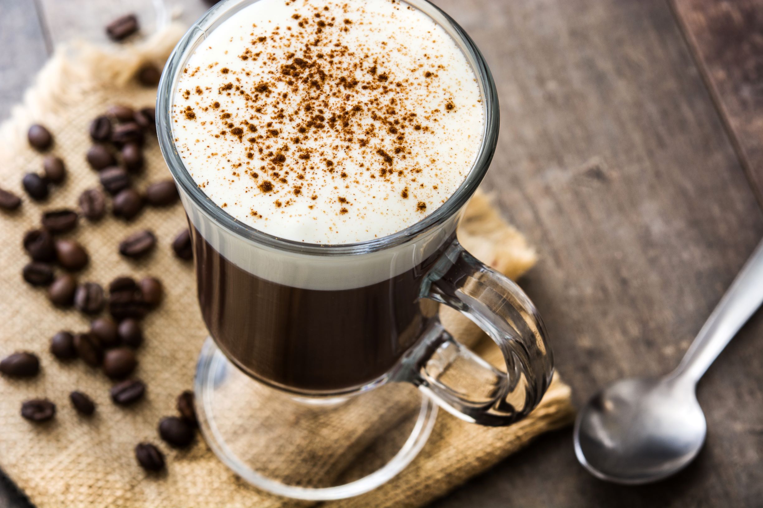 Cup of irish coffee in a glass mug sitting on a wooden table next to loose coffee beans and a spoon.