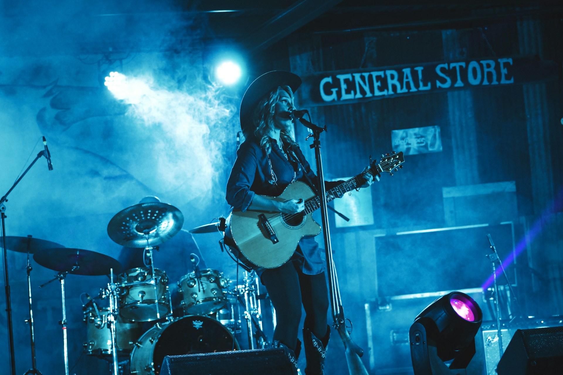 A female country singer playing guitar with her band on a blue lit stage.