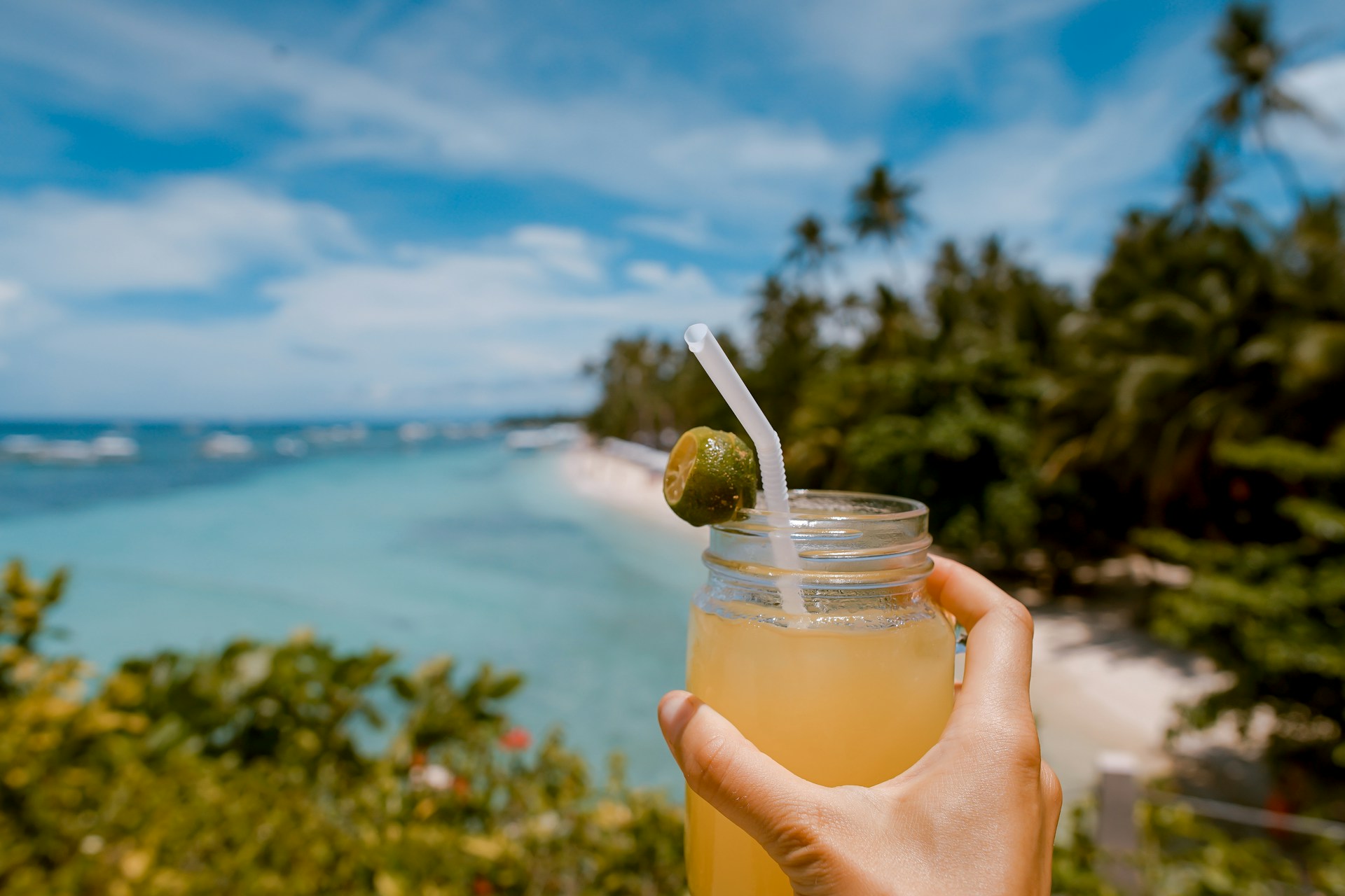 A hand holding a mason jar cocktail in front of a beach