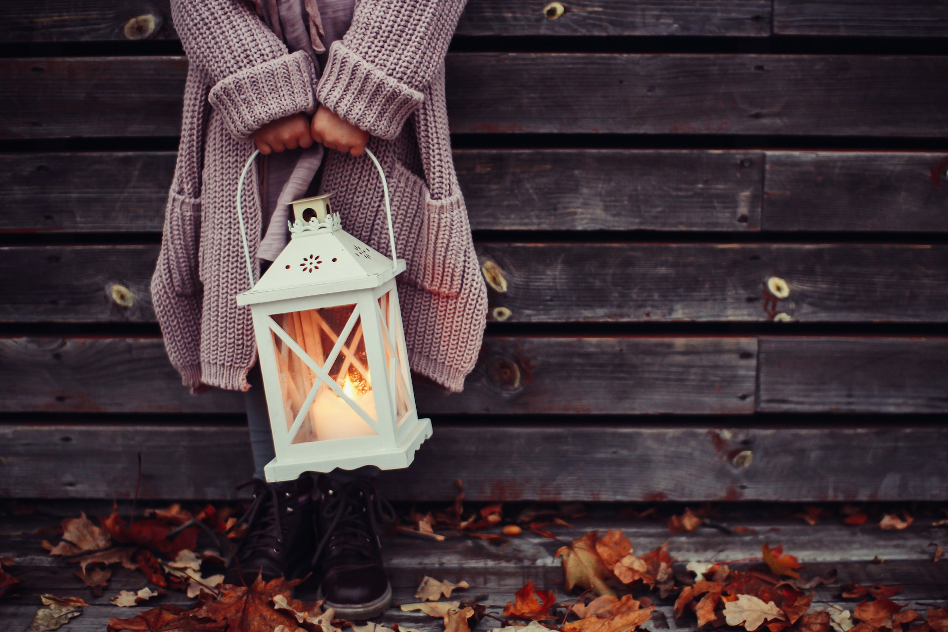 A woman's figure in a grey cardigan standing in front of a wooden building holding a lamp lit by a candle